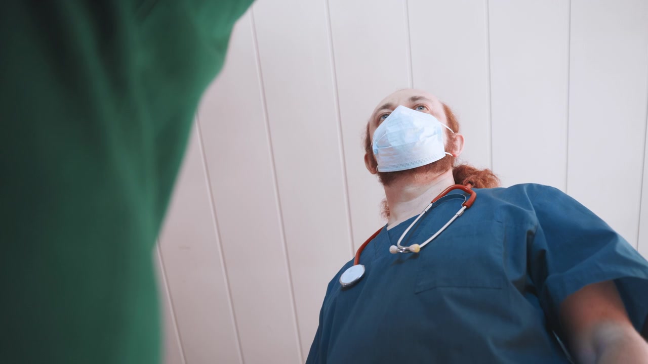 Medico di emergenza bianco a casa controlla i parametri vitali con lo stetoscopio, risposta mascherata, divisa blu, valutazione mirata vicino all'ingresso, urgenza clinica, equipaggiamento protettivo e attrezzatura visibili, supporto rapido