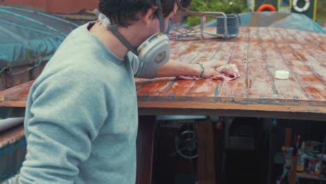 young man wipes boat wheelhouse roof planks with methylated spirits before varnishing