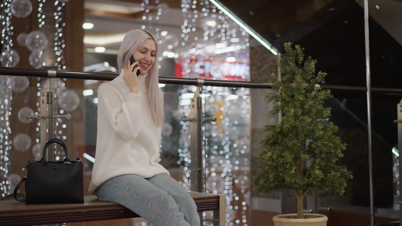 browsing shopper seated on bench in mall laughs during phone call, lifts leg playfully, white sweater and jeans, black bag at side, glowing festive lights and massage chairs in background
