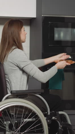 young woman with injury moves about kitchen in wheelchair. female person heats up food putting plate into microwave and waits calmly side view