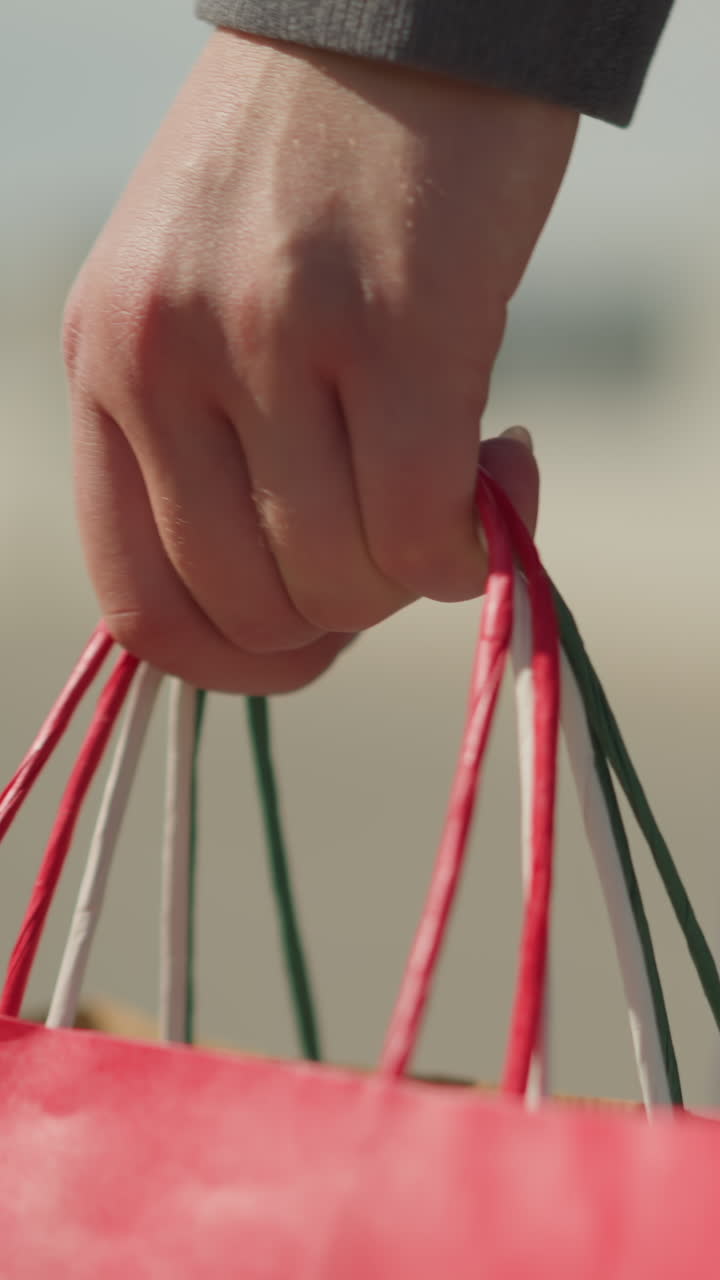 primer plano de un individuo con ropa gris sosteniendo bolsas de compras coloridas mientras camina, la mano agarra bolsas rojas y verdes, con un fondo borroso que da enfoque a la acción de llevar las bolsas
