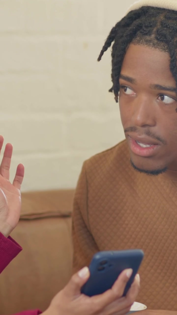 Vertical video: Reading man on tan couch scrolling blue phone with white cup, turning to maroon arm