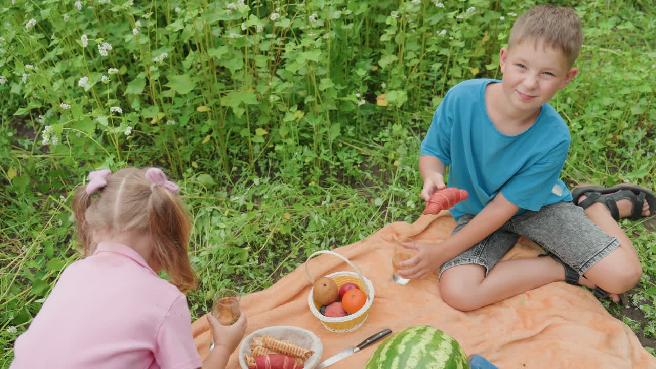 Un pícnic al aire libre con dos niños blancos; un niño con una camiseta verde azulado le ofrece una rodaja de sandía a una niña con coletas sobre una manta naranja; una cesta llena de manzanas y melón; un exuberante fondo de césped; verano natural