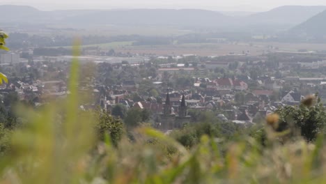 Vista-Borrosa-De-Una-Ciudad-Europea-Con-Chapiteles,-Enclavada-En-Un-Valle,-Desde-Un-Primer-Plano-Borroso-De-Hierba