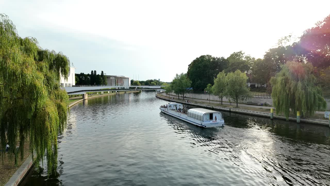 A tour boat cruising on a river flanked by trees and urban architecture