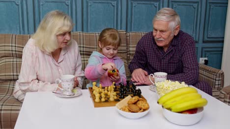 Senior-grandparents-and-child-granddaughter-spending-time-home-together,-sitting,-playing-chess-game