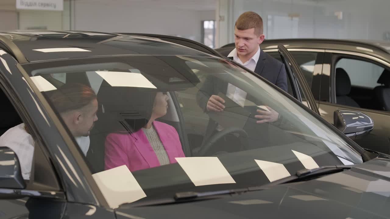 un joven vendedor de coches mostrando a una pareja de jóvenes un nuevo automóvil en el salón de concesionarios.