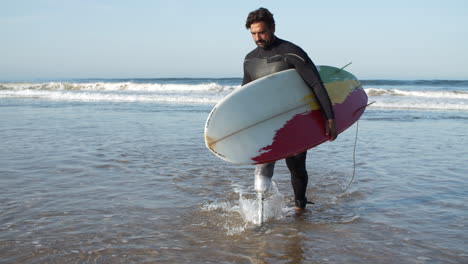 front view of a serious surfer with bionic leg coming out of ocean holding surfboard under arm 1