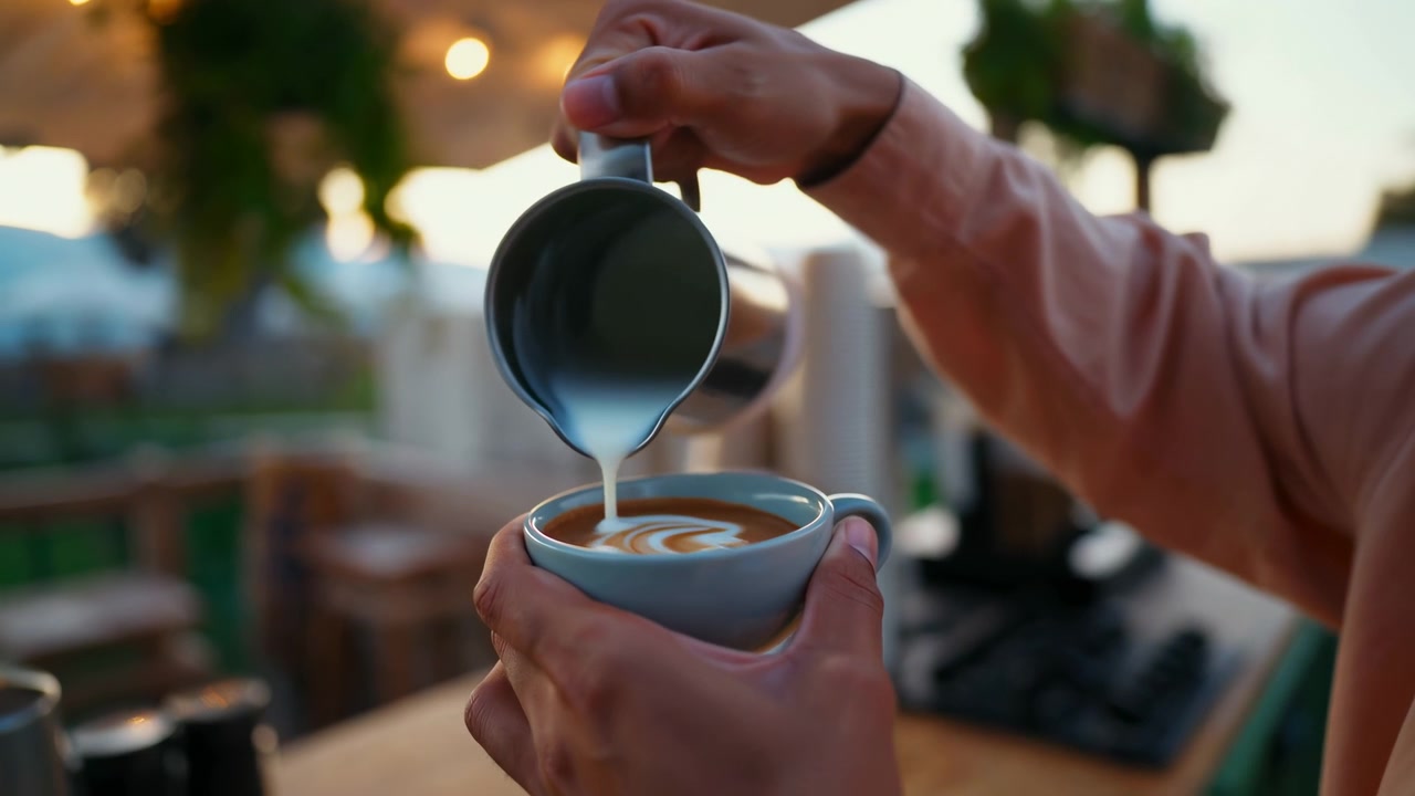 Un flusso costante di latte montato a vapore scorre nel caffè mentre si sviluppa il disegno sulla superficie, mentre luci calde sospese, arredi in legno e uno sfocato leggero dello sfondo incorniciano la vista ravvicinata.
