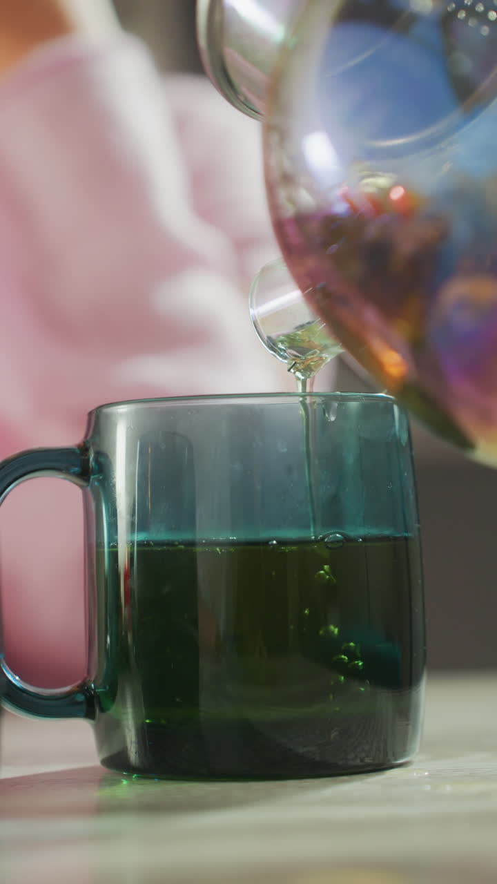 Tè verde che viene versato in una tazza di vetro, un calmo rituale di benessere casalingo con mani che inclinano una teiera iridescente per riempire la tazza trasparente, vapore leggero che sale, infusione di erbe che si deposita, sfondo bokeh morbido.