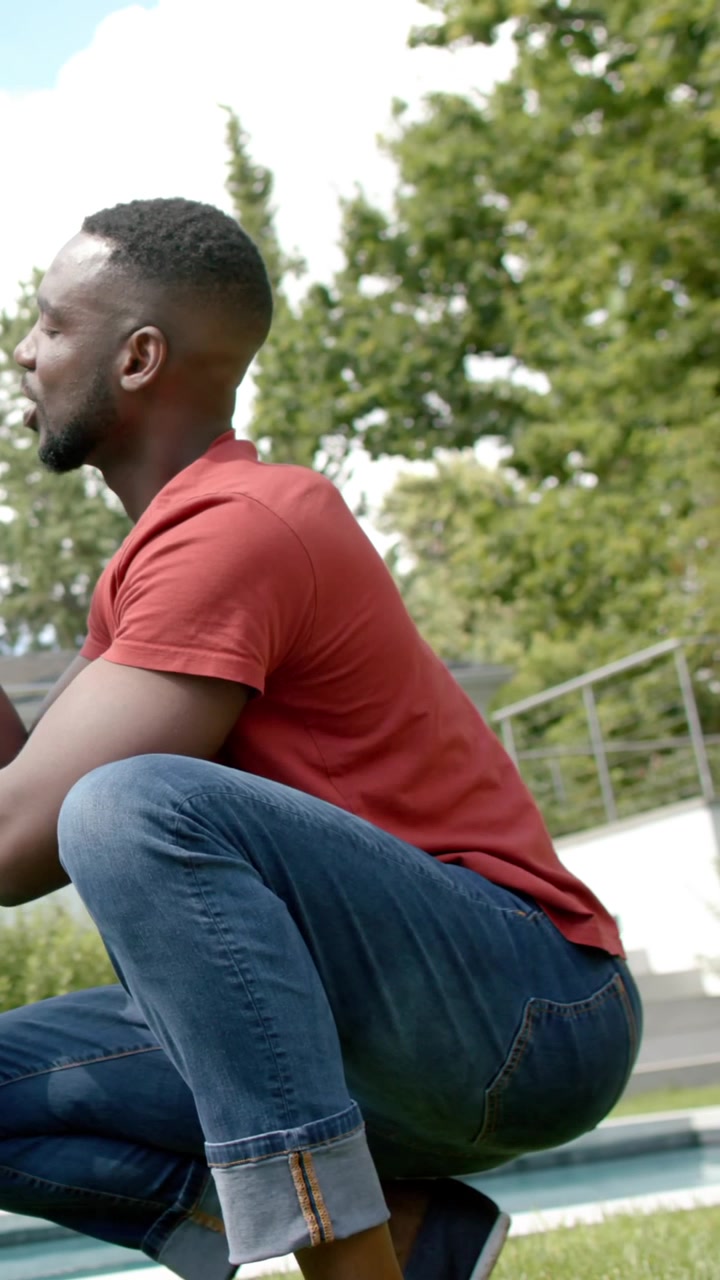 Vertical video: Crouching African American man on lawn by pool, shifting and smiling in red tee