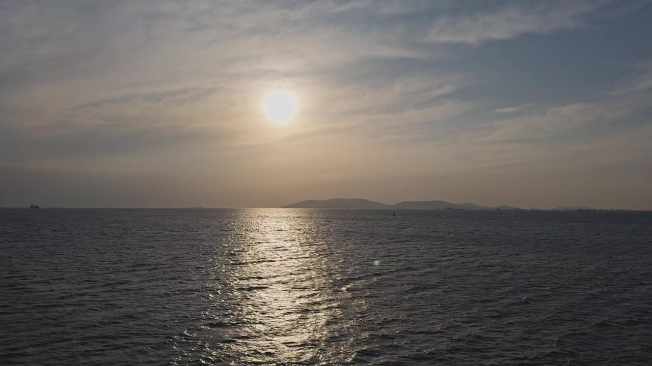 Static wide shot of a hazy golden sunset over the calm ocean horizon, with sunlight reflecting on the rippling water surface under a cloudy sky.