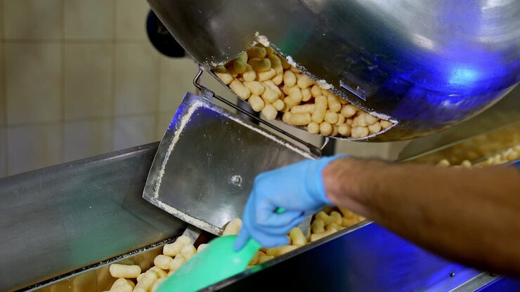 Sweet yellow crispy snacks falling on the conveyor line from opened metal tank. Maize sticks production process. Close up.