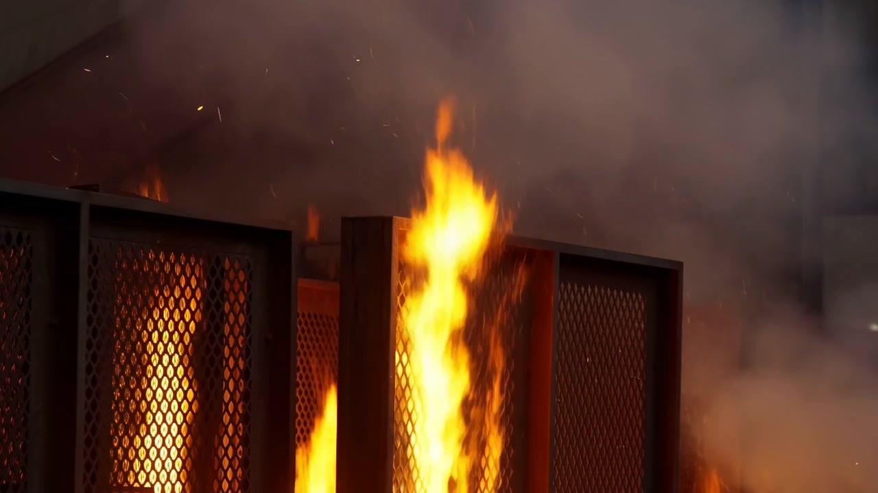 Bright orange flames surge upward through diamond pattern steel mesh barrier, sending thick smoke, glowing embers, and sparks into dark industrial space with dramatic high contrast light