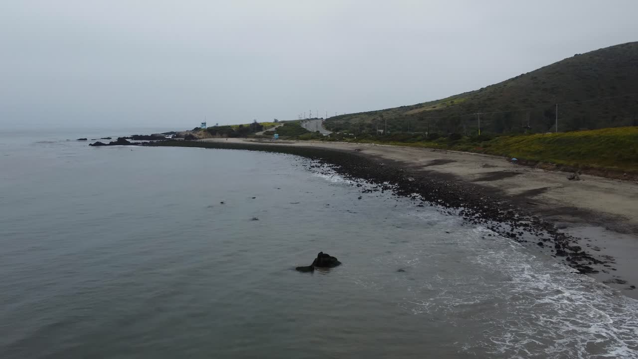 Low aerial shot of waves rolling over black rocks on a foggy beach on the Southern California coast. The road and lush green hills highlight the background as the ocean dominates the foreground.