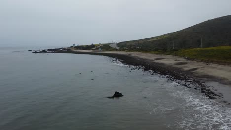low aerial shot of waves rolling over black rocks on a foggy beach on the southern california coast. the road and lush green hills highlight the background as the ocean dominates the foreground.