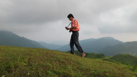 Shot-of-an-Asian-man-musician-walking-on-grass-on-top-on-a-mountain-and-playing-ukulele-on-a-rainy-day