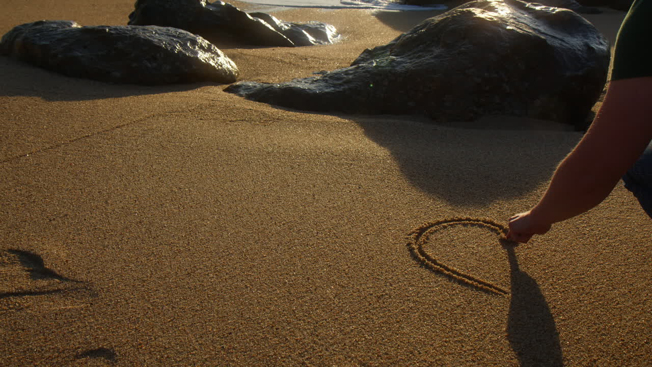 Romantic boy drawing a heart in the sand of a beach. Slow motion