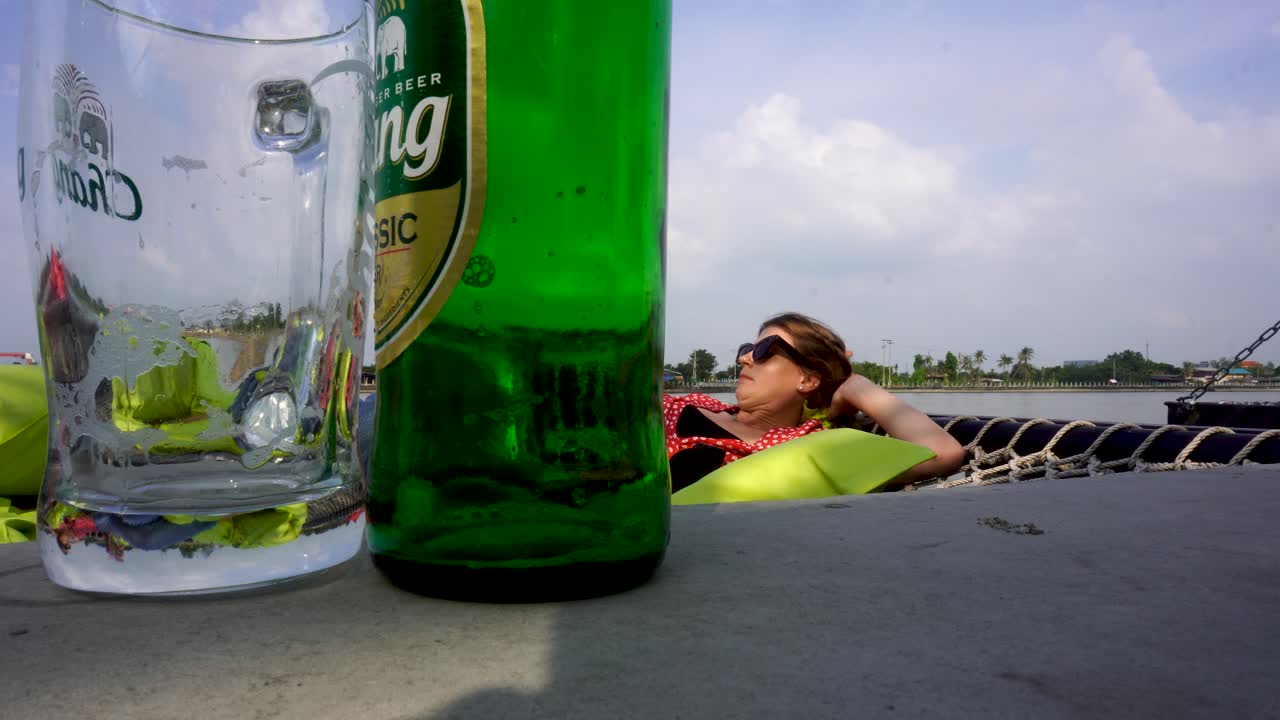 Close Up View Of Empty Beer Bottle With Glass On Table With Female Tourist Relaxing On Hammock In Background. Locked Off