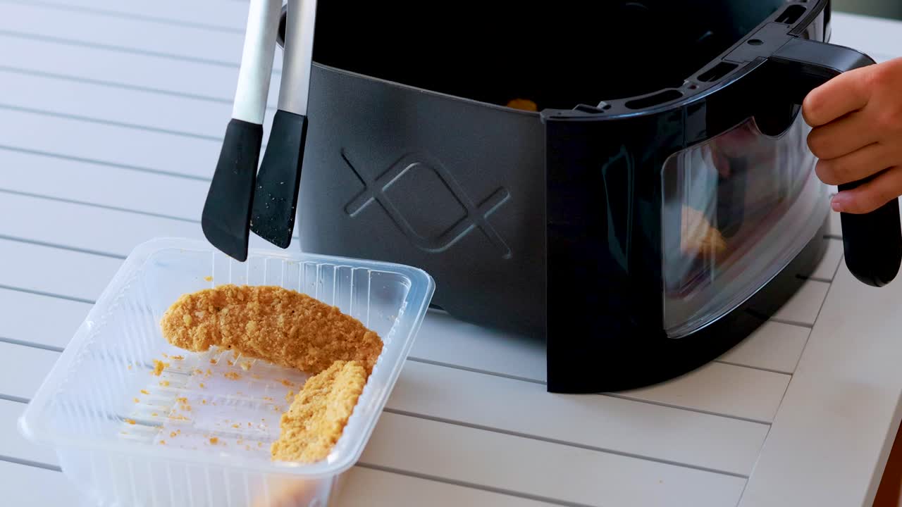 A person uses metal tongs to pick up breaded food from a plastic tray and place it into a black air fryer on a white table in bright, natural lighting