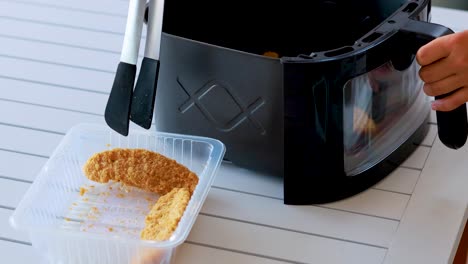 a person uses metal tongs to pick up breaded food from a plastic tray and place it into a black air fryer on a white table in bright, natural lighting