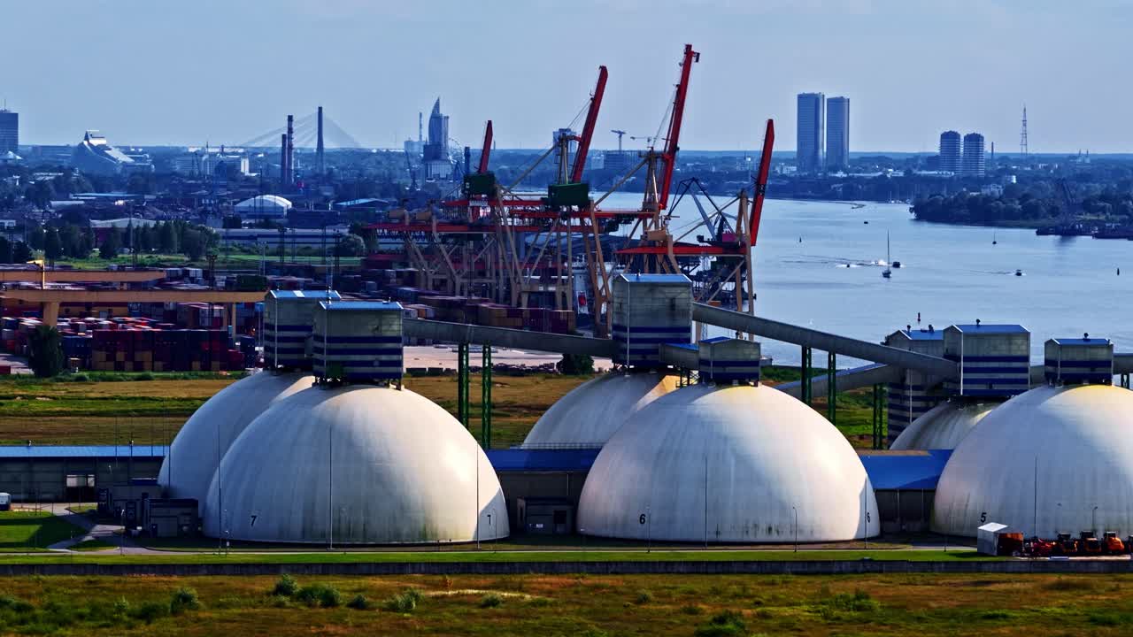 Vue aérienne de grands dômes de stockage blancs près d’un port fluvial animé, avec des grues, des conteneurs et une ligne d’horizon urbaine lointaine formant un paysage industriel