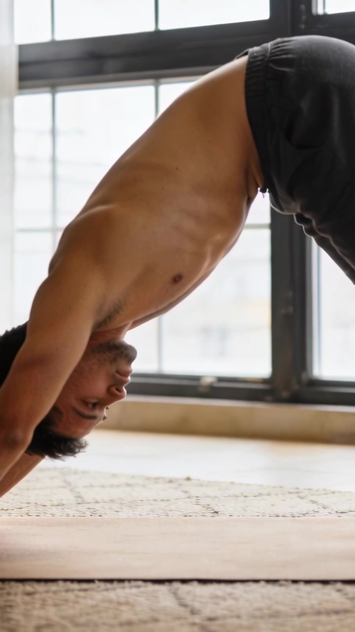Video verticale: Uomo a torso nudo che inizia con le mani a terra, mantenendo la posizione del cane a testa in giù a casa su un tappetino per fare stretching.
