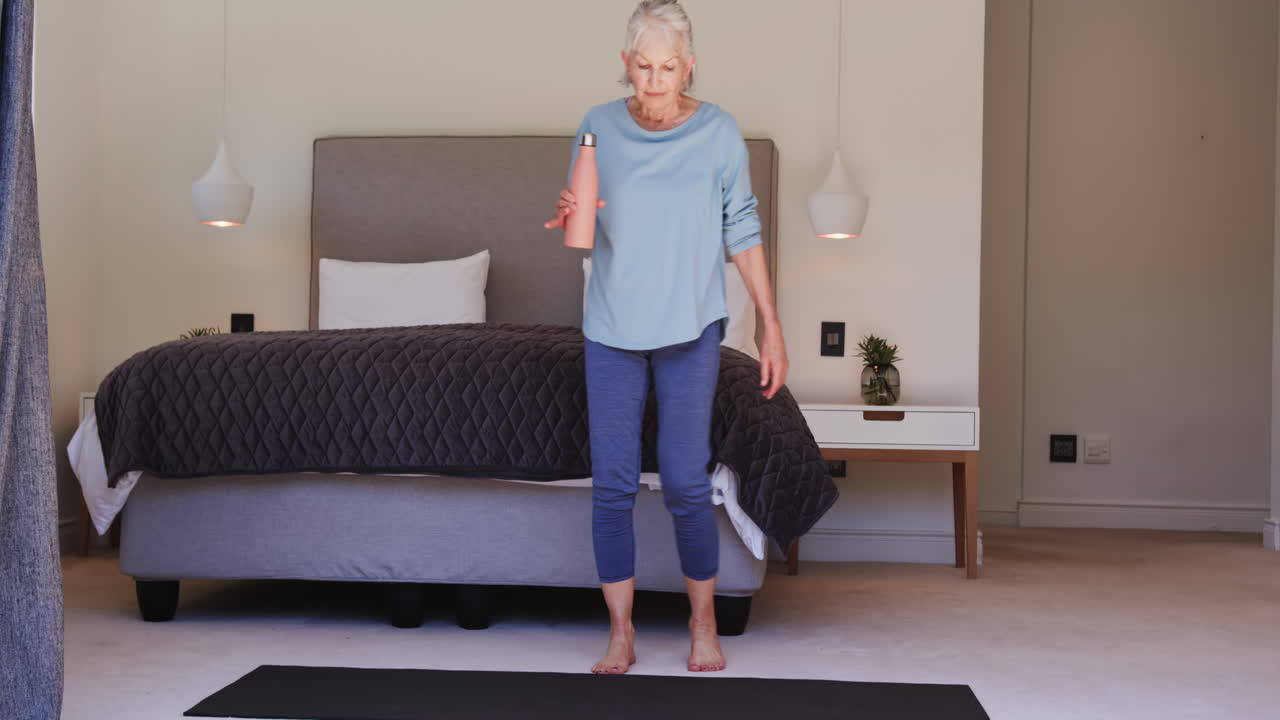 Senior woman relaxing on yoga mat at home, holding water bottle
