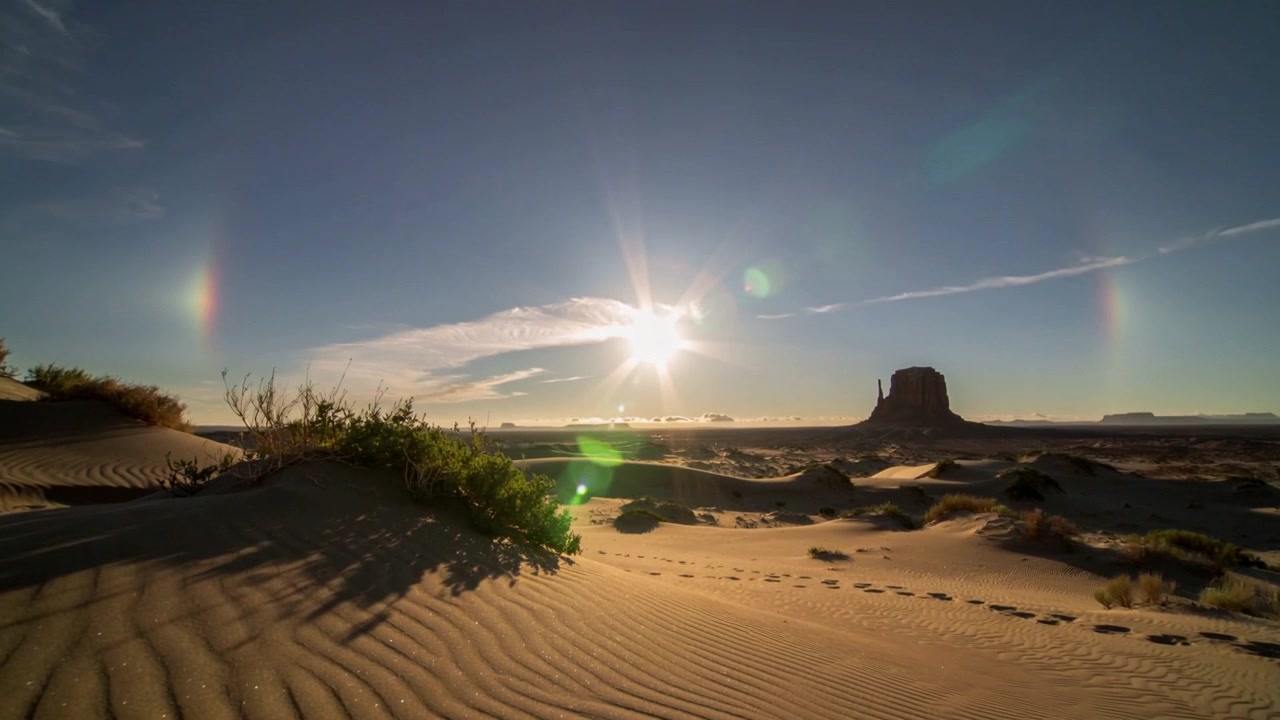 Sol nascente iluminando duna em primeiro plano ao nascer do sol no deserto, formando parélios e revelando pegadas.