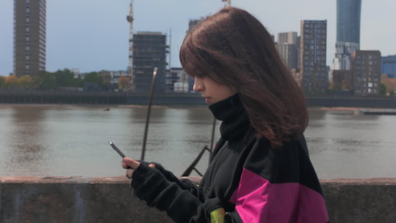Vista laterale di una giovane donna che cammina vicino a un fiume in città mentre usa il suo cellulare.