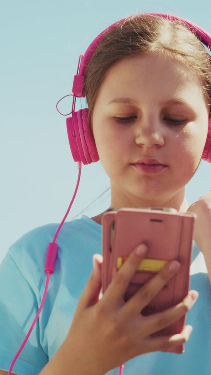 Bright pink headphones on young girl's ears. She holds a phone, eyes closed, head tilted. Soft sunlight glows behind her, gently. Focused on music, she seems peaceful. Ideal for youth, relaxation
