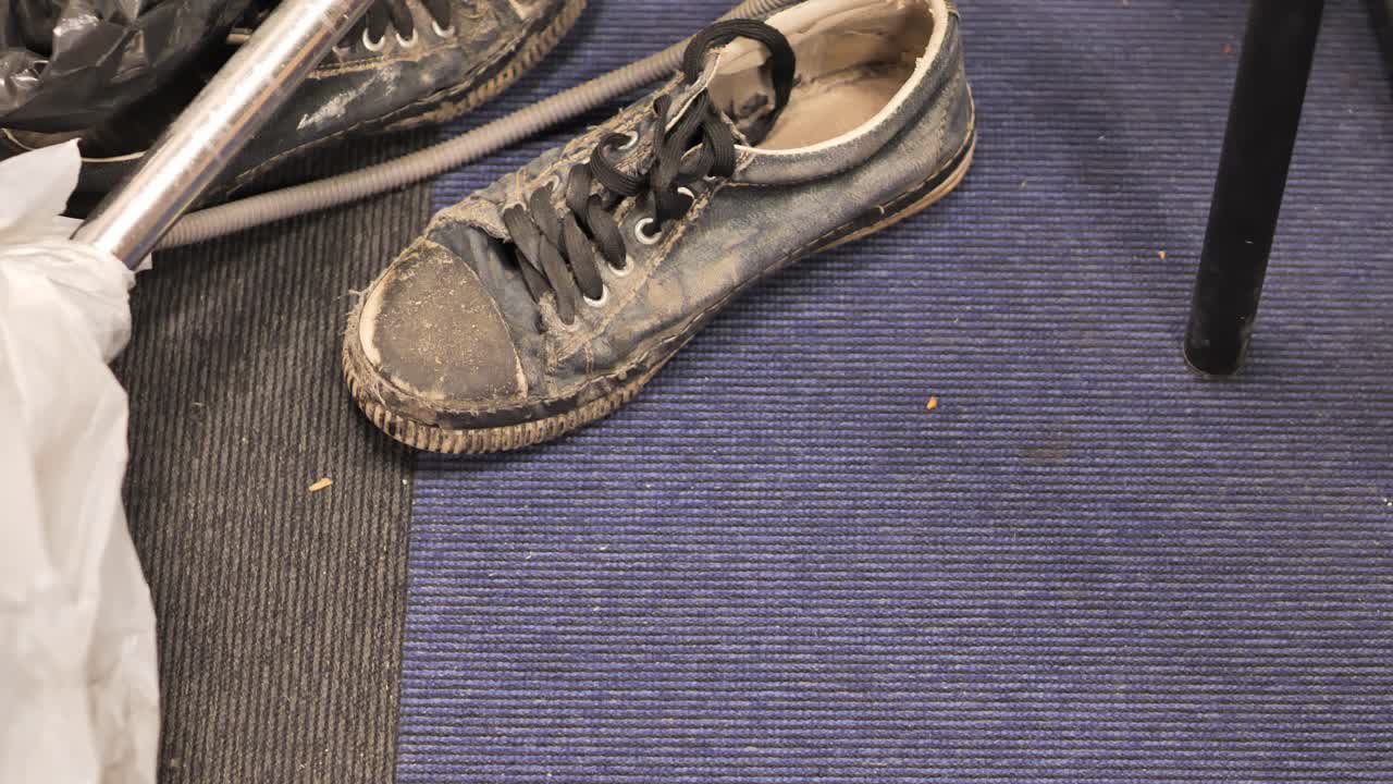 Camera pans from dirty shoe on carpet to grass trimmer, showing messy workspace.