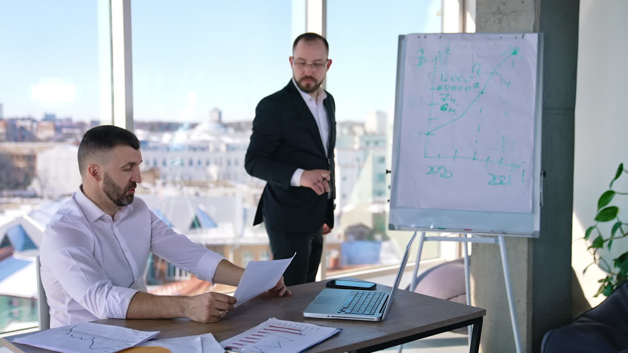 Two businessmen discussing something in a light office. One man sitting at the table and holding a paper. Business partner standing at the whiteboard looking at the chart and commenting it.