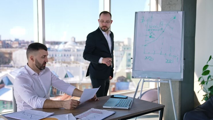 Two businessmen discussing something in a light office. One man sitting at the table and holding a paper. Business partner standing at the whiteboard looking at the chart and commenting it.