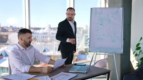 two businessmen discussing something in a light office. one man sitting at the table and holding a paper. business partner standing at the whiteboard looking at the chart and commenting it.