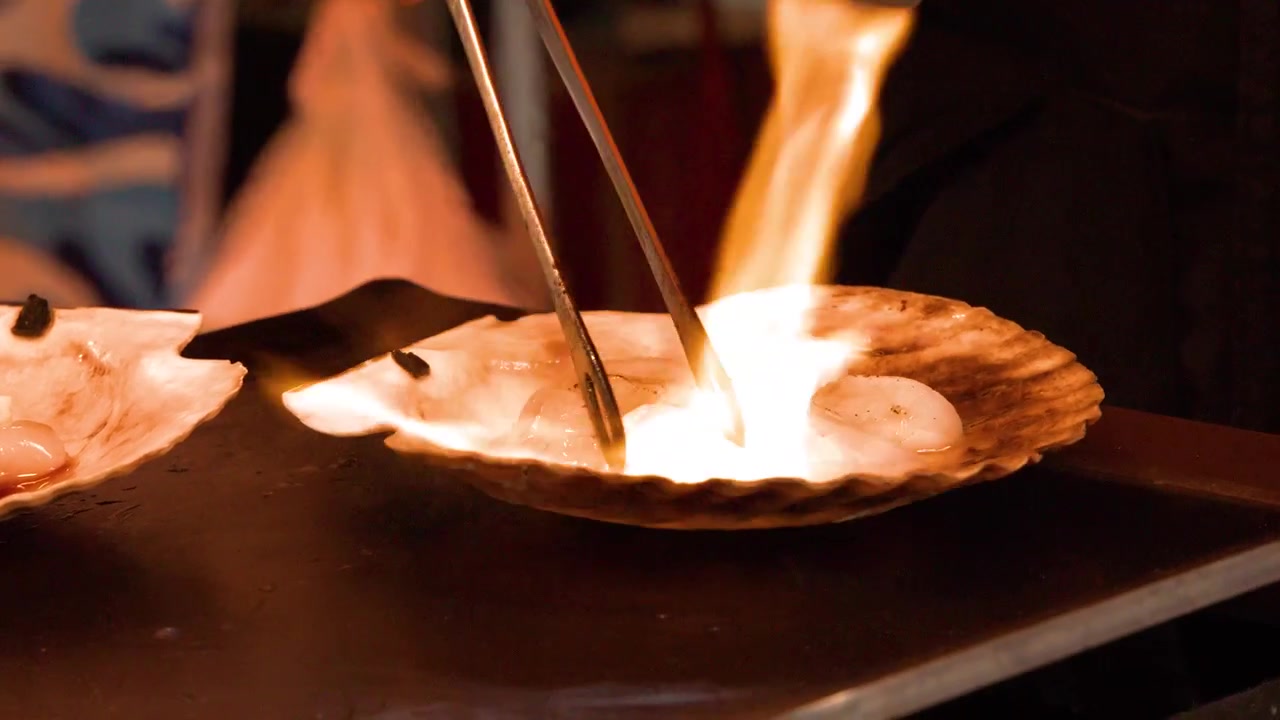 Close-up shot of a street food vendor using a blowtorch to sear scallops in shells