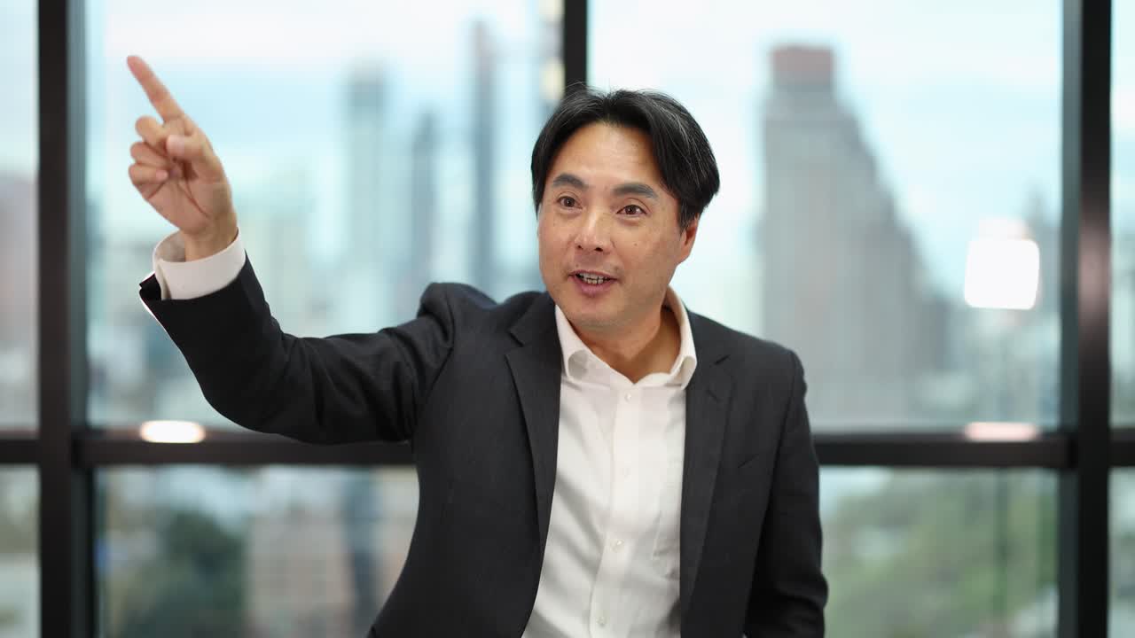Confident Asian businessman gestures and speaks energetically in bright office with cityscape background