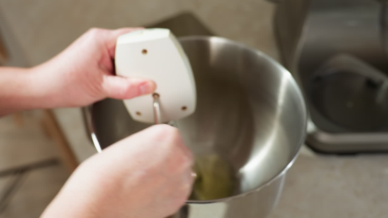 Close up of cook using electric hand mixer to whisk raw egg mixture inside stainless steel bowl on kitchen counter, showing motion and focus on blending process with firm grip on mixer handle