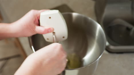 close up of cook using electric hand mixer to whisk raw egg mixture inside stainless steel bowl on kitchen counter, showing motion and focus on blending process with firm grip on mixer handle