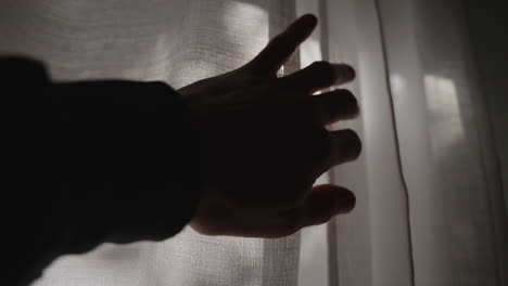 hand reaching towards white window curtain - opening fabric to let sun shine inside dark room