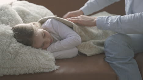 little daughter sleeps in the sofa, her dad covers her with a quilt