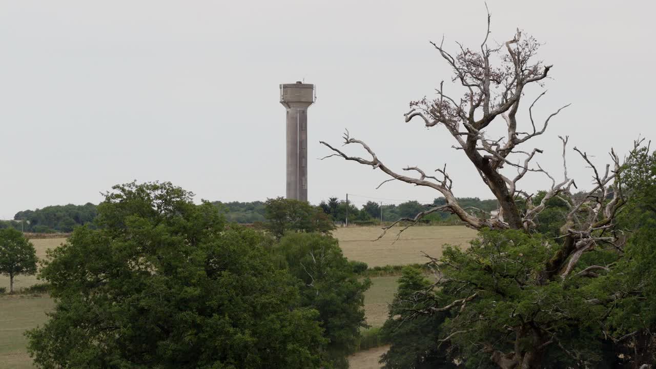 A tall concrete water tower stands in the countryside, surrounded by fields and trees, captured in a wide aerial shot