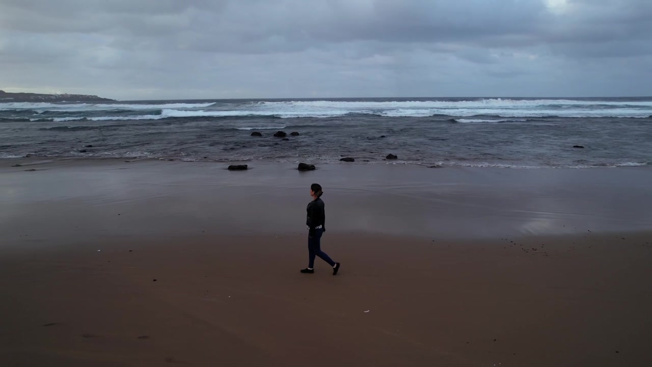 Donna pensierosa che cammina da sola lungo la riva di una spiaggia desolata in una giornata nuvolosa. Una scena toccante che trasmette solitudine e introspezione