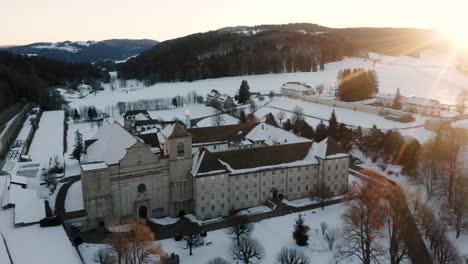 The-abbey-of-Bellelay,-historical-monument-of-the-Bernese-Jura-in-Switzerland,-drone-view-during-a-cold-winter-and-snowy-sunrise