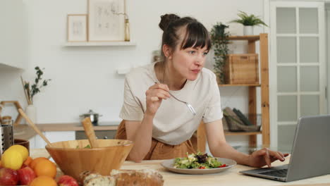 young woman eating salad and using laptop in kitchen