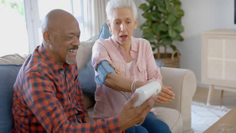 Feliz-Pareja-Birracial-Mayor-Sentada-En-El-Sofá-Y-Tomando-Presión-En-Casa,-Cámara-Lenta