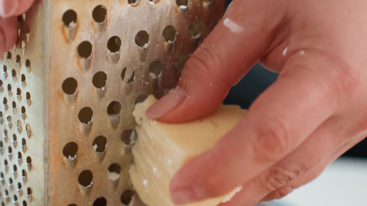 Hands actively grating soft cheese against metal box grater, creating small shavings that collect in perforated holes and cling to fingertips during focused kitchen preparation, with cheese, grater