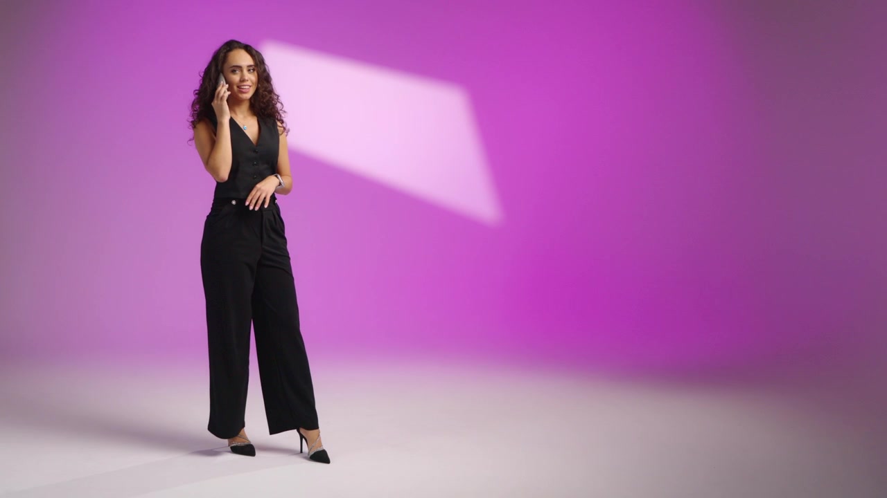 Donna elegante con capelli ricci entra nell'inquadratura su uno sfondo viola da studio, indossando tacchi neri e un completo, sorridendo mentre parla al telefono e facendo gesti espressivi con le mani