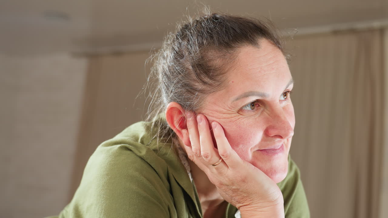Thoughtful woman resting head on hand with eyes gently closed and warm smile, dressed in green kitchen attire, radiating calm and contentment in cozy home setting filled