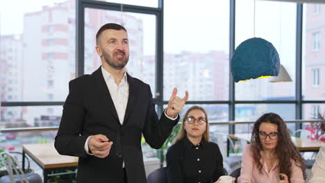 Bearded businessman in black suit speaks to the camera. Office team sitting at the table beside listening to boss.